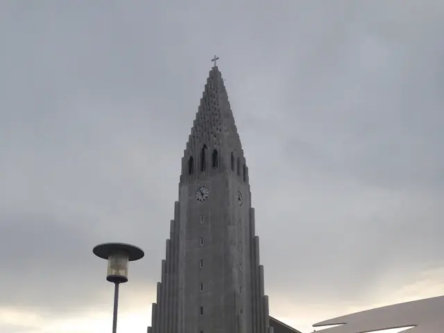 In this image there is a building with clock on it, also there are some trees and electrical pole...