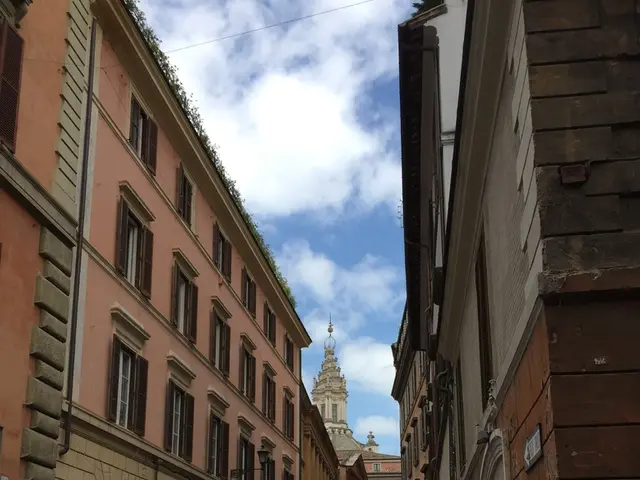 In this image there are buildings, plants and there are vehicles on the road and the sky is cloudy.